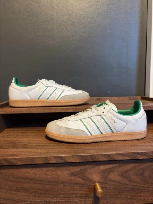 Adidas Samba vita och gröna sneakers - Klassiska Adidas Samba sneakers i vitt läder med gröna detaljer och gummisula. Skorna har de ikoniska tre ränderna på sidan, grönt foder och logga på plösen. Tåpartiet är i ljusgrå mocka och yttersulan är i naturfärgat gummi. Perfekta för en sportig och stilren look.