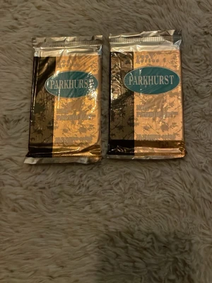 Parkhurst samlarkortpaket 1991 - Två oöppnade paket med Parkhurst samlarkort från 1991. Förpackningarna är i guld och svart folie med blå Parkhurst-logga. Perfekt för dig som gillar att samla på sportkort eller vill ha något unikt från 90-talet.