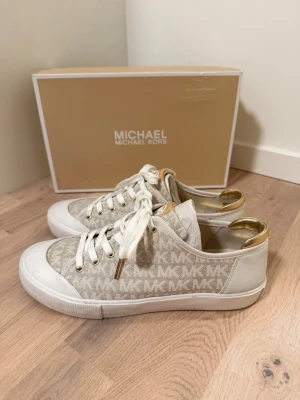 Beige MK sneakers med gulddetaljer - Snygga sneakers från Michael Kors i beige med MK-logga över hela skon, vita snören och gulddetaljer vid hälen. Ovandelen är i textil och syntet, sulan är vit och tåpartiet har en klassisk gummidetalj. Perfekta för dig som gillar stilrena och trendiga skor 🤍känns som 40