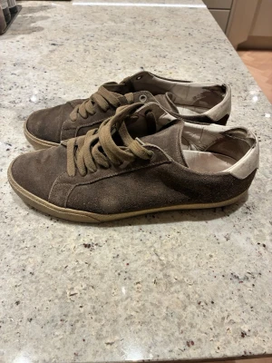 Bruna Saint laurent mocka sneakers - Snygga bruna sneakers i mocka med beige detaljer på hälen och matchande bruna skosnören. Låg modell med klassisk siluett och gummisula. Perfekta för en avslappnad och stilren look. Bruksskick, använda.