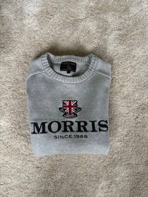Grå tröja från Morris Gents - Snygg grå tröja från Morris Gents med broderad logga och brittisk flagga på bröstet. Rund halsringning och ribbade muddar. Perfekt för en stilren och klassisk look.