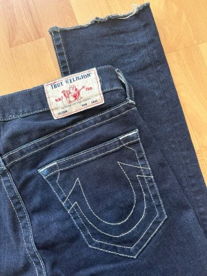 True Religion jeans - Mörkblå jeans från True Religion med ikonisk kontrastsöm och stor logga på bakfickan. Raka ben med rå, fransad kant nedtill. Klassisk femficksmodell i denim med tydliga vita sömmar. Storlek 32💫
