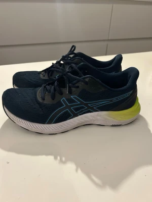 Mörkblå Asics sneakers med limegrön detalj - Snygga mörkblå sneakers från Asics med ventilerande mesh-ovandel, svarta och blå detaljer samt limegrön häl. Skorna har klassisk snörning och en vit, dämpad sula för extra komfort. Perfekta för dig som gillar sportig stil och vill ha bekväma skor till vardagen.