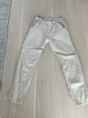 Beige linnebyxor  - Snygga beige linnebyxor med raka ben. Byxorna har två fickor fram och är tillverkade i ett lätt och luftigt linnematerial, perfekta för en avslappnad och stilren look. Linder 164 cm storlek