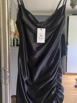 Helt NY med lapp! Svart satin klänning från Only - Svart kort klänning i glansigt satinmaterial från Only. Klänningen har tunna axelband, draperad halsringning och rynkade detaljer längs ena sidan för en snygg siluett. Något stretchig. Perfekt för dig som vill ha en stilren och trendig look.
