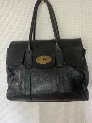 Svart Mulberry Bayswater handväska - Klassisk svart Bayswater handväska från Mulberry i äkta läder. Väskan har dubbla handtag, vridlås i mässing och en stilren, rymlig design. Perfekt för dig som gillar tidlös och lyxig stil. Ikonisk metallplatta framtill med Mulberry-logga.