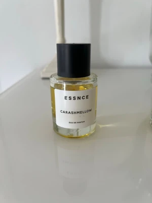ESSNCE Carashmellow Eau de Parfum - En stilren parfymflaska från ESSNCE med doften Carashmellow. Flaskan är cylindrisk i genomskinligt glas med svart matt kork och vit etikett med svart text. Innehållet är ljusgult och ger ett modernt och minimalistiskt intryck.