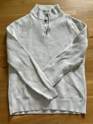 Beige stickad tröja med halvzip - Snygg beige stickad tröja från These Glory Days med halvzip och hög krage. Tröjan har ett diskret rutmönster i stickningen, långa ärmar och ribbade muddar. Perfekt för lager-på-lager och enkel att matcha med jeans eller chinos.
