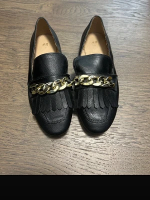 Svarta loafers med guldkedja H&M - Stilrena svarta loafers från H&M med dekorativ guldfärgad kedja och fransdetaljer framtill. Skorna har en klassisk, rundad tå och är tillverkade i konstläder med slät finish. Perfekta för dig som vill ha en trendig och snygg look.
