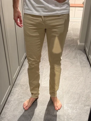 Beige jeans från J.Lindeberg - Snygga beige jeans från J.Lindeberg med klassisk femficksmodell och raka ben. Jeansen har en stilren design med läderpatch bak i midjan och silverfärgade nitar. Perfekta för dig som vill ha en clean och modern look. Modell: Jay Storlek: 31/34
