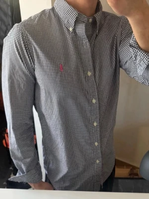 Rutig skjorta från Ralph Lauren - Klassisk rutig skjorta från Ralph Lauren i svart och vitt med knappkrage och lång ärm. Skjortan har en liten rosa logga broderad på bröstet och är tillverkad i bomull. Perfekt för en stilren och avslappnad look.