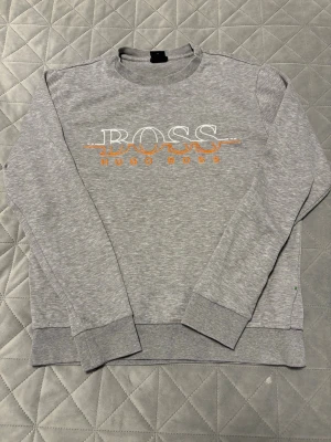 Grå sweatshirt från Hugo Boss - Grå sweatshirt från Hugo Boss med broderad logga i vitt och orange framtill. Tröjan har rund hals, långa ärmar och ribbade muddar vid ärmslut och nederkant. Perfekt för en chill och stilren vibe.