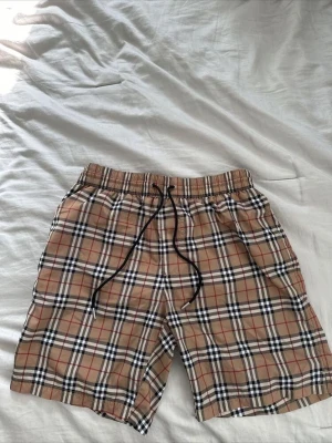 Beige rutiga shorts med snörning - Säljer ett par klassiska beige shorts från Burberry med ikoniskt rutigt mönster i svart, vitt och rött. Shortsen har elastisk midja och svart snörning framtill. Perfekta för sommaren och ger en stilren vibe till din outfit.
