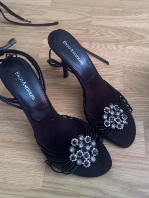 Svarta klackar Enzo Angiolini - Snygga svarta pumps med smala remmar och öppen tå från Enzo Angiolini. Skorna har en elegant klack och är dekorerade med stora blommor av strass framtill. Perfekta för dig som vill sticka ut med en glittrig detalj.