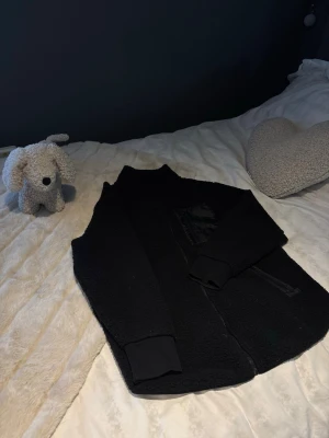 Svart teddyjacka med dragkedja - Mysig svart teddyjacka med hel dragkedja framtill och två sidofickor med dragkedja. Bröstficka med detaljer i nylon ger en cool kontrast. Jackan har hög krage och muddar vid ärmsluten. Perfekt för kalla dagar och enkel att styla.
