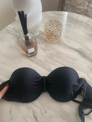 Svart bandeau-bh med knytband - Snygg svart bandeau-bh med vadderade kupor och knytband i ryggen. Bh:n har avtagbara axelband och en enkel rosett mellan kuporna. Perfekt för axellösa toppar och klänningar. Materialet känns mjukt och stretchigt.
