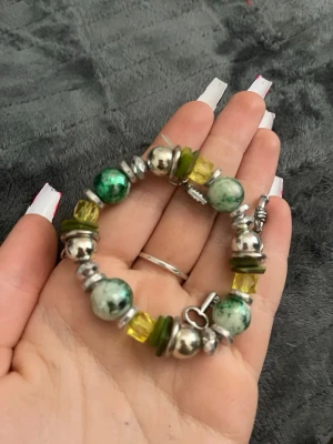 Grönt och silverfärgat pärlarmband - (Pris kan diskuteras)Snyggt armband med gröna, gula och silverfärgade pärlor i olika former. Armbandet har detaljer i metall och ett dekorativt lås. Perfekt accessoar för att ge din outfit en färgklick och unik stil.