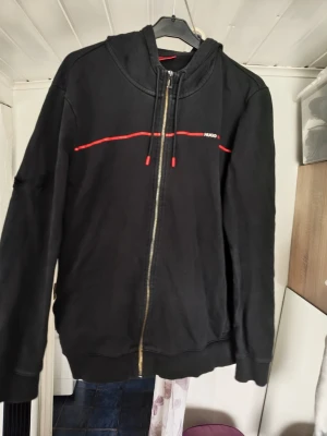Svart hoodie med dragkedja från HUGO - Svart hoodie från HUGO med dragkedja framtill, röda detaljer och dragsko i huvan. Snygg röd linje över bröstet och diskret HUGO-logga. Perfekt för en stilren och avslappnad look. Hål i den samt något märke 