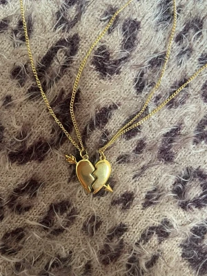 Guldiga best friends-halsband hjärta - Två matchande halsband i guldton med varsin halva av ett trasigt hjärta som hänge. Ena halvan har en pil och den andra en krona, perfekt för att dela med en vän. Kedjorna är tunna och glänsande, och hjärtana har en slät, blank yta. Rostfri 