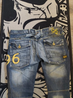 G-star Blå jeans med gula detaljer, 96-print - Snygga G-star blå jeans med slitningar och stora bakfickor. Gula detaljer och tryck med siffran 96 på ena benet och bakfickan. Raka ben och dekorativa sömmar. Perfekta för dig som gillar streetwear och vill sticka ut med coola detaljer.