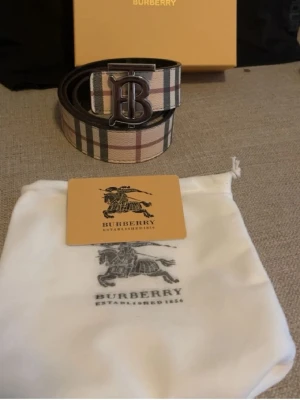 Burberry rutigt bälte med logga - Snyggt bälte från Burberry med klassiskt rutigt mönster i beige, svart och rött. Bältet har ett stort metallspänne med Burberrys ikoniska 'B'-logga. Perfekt accessoar för att lyfta din outfit.