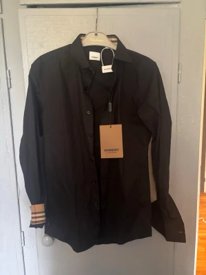 Svart skjorta från Burberry - Stilren svart skjorta från Burberry med klassiska rutiga detaljer på insidan av manschetterna och kragen. Skjortan har knappar framtill och lång ärm. Perfekt för dig som gillar exklusiva märken och snygga detaljer.