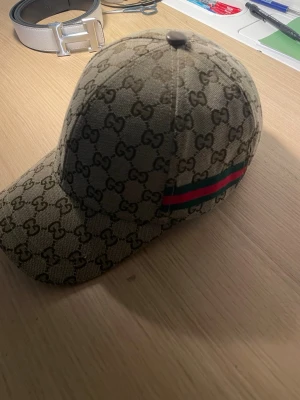 Gucci keps med GG-mönster - ”Kepsen är i väldigt bra skick, vinkeln som bilden togs ifrån och ljuset gjorde så att den såg lila ut” Snygg keps från Gucci i beige canvas med klassiskt GG-monogram över hela kepsen. På sidan finns den ikoniska gröna och röda randen. Perfekt accessoar för dig som vill sticka ut med lyxig streetstyle.