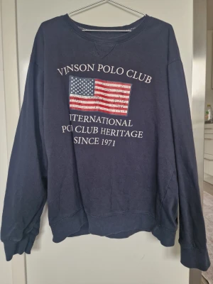 Marinblå sweatshirt Vinson Polo Club - Mysig marinblå sweatshirt från Vinson Polo Club med amerikansk flagga och vit text på bröstet. Tröjan har rund halsringning, långa ärmar och ribbade muddar. Perfekt för en avslappnad och sportig stil. Är xl storlek men sitter som L nästan