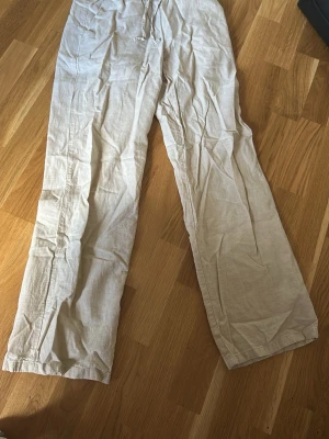 Beiga linnebyxor med dragsko - Snygga och luftiga beiga linnebyxor med rak passform och dragsko i midjan. Perfekta för en avslappnad stil och tillverkade i ett lätt linnematerial som andas. Byxorna har en enkel design utan synliga fickor eller detaljer.