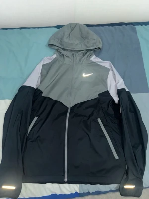 Nike vindjacka grå - Nike vindjacka med luva i grått och svart med vita detaljer. Jackan har hel dragkedja, två fickor med dragkedja och reflekterande detaljer på armar och bröst. Perfekt för blåsiga dagar och sportiga outfits.