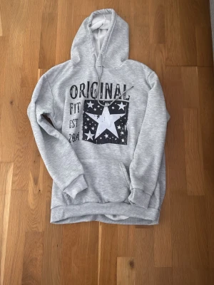 Grå hoodie med stjärntryck - Säljer en grå hoodie från Original Fit med stor vit stjärna och svart tryck med text på bröstet. Hoodien har huva med dragsko, ribbade muddar och en klassisk passform. Perfekt för dig som gillar en avslappnad och cool stil.