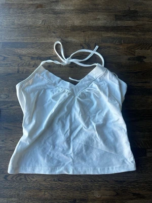 Vit halterneck topp - Säljer en vit halterneck topp med knytband i nacken och v-ringning. Toppen har en enkel och stilren design. Perfekt till sommaren och enkel att matcha med olika outfits.