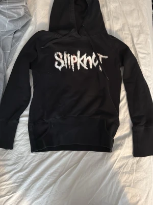 Svart Slipknot hoodie med tryck - Svart hoodie med Slipknot-logga i vitt på bröstet och ett stort grafiskt tryck på ryggen. Klassisk huva med magficka framtill. Perfekt för dig som gillar bandmerch och vill ha en streetwear-look. Bra skick, storlek S. Säljer då ja inte lyssnar på slipknot längre.
