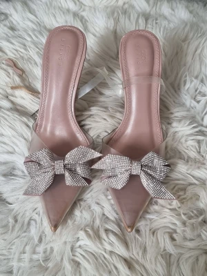 Rosa pumps med strassrosett från ASOS - Snygga pumps i ljusrosa med spetsig tå och transparenta detaljer. Skorna har en hög klack och är dekorerade med en stor glittrig strassrosett framtill. Perfekta för dig som vill sticka ut med en trendig och glamorös look.