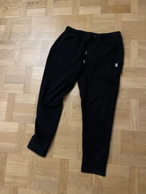 Svarta mjukisbyxor från Polo Ralph Lauren - Svarta mjukisbyxor från Polo Ralph Lauren med klassisk broderad logga på benet. Byxorna har dragsko i midjan och muddar vid bensluten. Perfekta för en avslappnad och stilren look. Tillverkade i mjukt material för extra komfort. Storlek L men passar mer M
