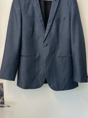 Mörkgrå klassisk kavaj herr - Stilren mörkgrå kavaj med enkelknäppning och smal passform. Kavajen har två framfickor, bröstficka och klassisk krage. Perfekt för en dressad look. Materialet ser ut att vara en blandning av ull och polyester.