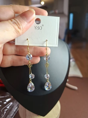Långa örhängen med kristallpärlor - Snygga långa örhängen från Y80 med guldtonad metall och flera klara kristallpärlor i rund och droppform. Örhängena har dekorativa små blommor i metall och ger ett glittrande intryck. Perfekta för att lyfta din stil med lite extra bling.