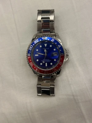 Arkench klocka med blå och röd bezel - Snygg klocka från Arkench med blå urtavla och datumvisning. Bezel i blått och rött ger en sportig look. Armbandet är i silverfärgat metall och har viklås. Perfekt accessoar för dig som gillar stilrena detaljer. Den är lute förminskad me men skulle passa dom flesta skruv vad du har för storlek så kan vi kolla om den passar 