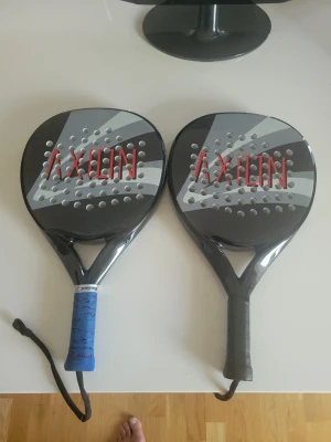 Två svarta padelracketar Axion - Säljer två svarta padelracketar från Axion med gråa och vita detaljer samt röd logga. Ena racketen har blått grepp och handledsrem, den andra har svart grepp och handledsrem. Perfekt för dig som vill spela padel med kompis.