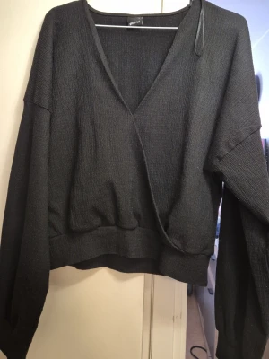 Svart omlottopp från Gina Tricot - Snygg svart omlottopp från Gina Tricot med djup V-ringning och vida ärmar. Tröjan har en ribbad struktur och resår i midjan för en skön passform. Perfekt att styla med jeans eller kjol för en trendig look.
