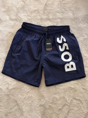 Marinblå badbyxor från BOSS strl S - Snygga marinblå badbyxor från BOSS med stort vitt logotryck på ena benet. Modellen har resår och dragsko i midjan samt sidofickor. Tillverkade i snabbtorkande material, perfekta för stranden eller poolen.