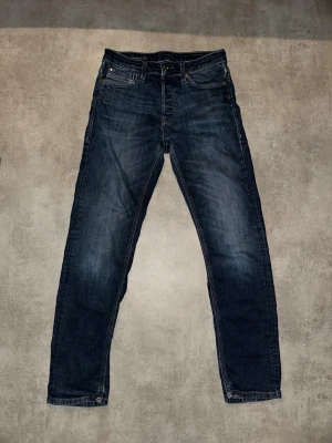 Mörkblå jeans från Jack and Jones  - Snygga mörkblå jeans från  med Comfort fit-passform. Klassisk femficksmodell med guldfärgade nitar och knapp. Jeansen har en lätt tvättad look och raka ben. Perfekta för en avslappnad och trendig stil. Hör av vid minsta fundering