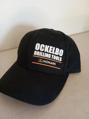 Svart keps Ockelbo Monark - Snygg svart keps med böjd skärm och broderad text 'OCKELBO DRILLING TOOLS' samt Monark-logga framtill. Kepsen har klassisk passform och justerbar rem bak. Perfekt för dig som gillar industriell stil och vill sticka ut.