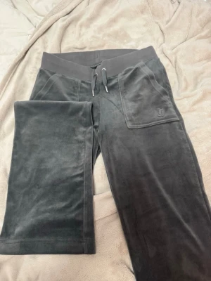 Mörkgrå velourbyxor Juicy Couture - Mörkgrå mjukisbyxor i velour från Juicy Couture med bred resår i midjan, dragsko och fickor fram. Bakficka med lock och tryckknappar. Perfekta för en chill och trendig look.