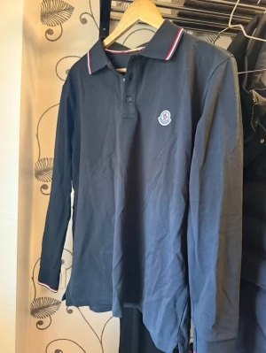 Svart långärmad piké från Moncler - Snygg långärmad pikétröja från Moncler med klassisk krage och knappar framtill. Tröjan har vita och röda detaljer på kragen och ärmsluten samt Moncler-logga på bröstet. Perfekt för en stilren och sportig look.