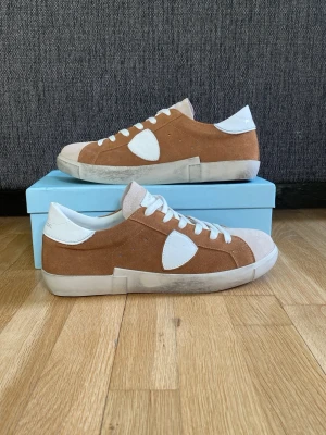 Philippe Modell skor - Snygga sneakers i mocka från Philippe modell. Skorna är bruna med vita detaljer, vita skosnören och vit sula. Klassisk låg modell med rund tå och diskret logga på hälen. Perfekta för dig som gillar stilrena och trendiga sneakers.