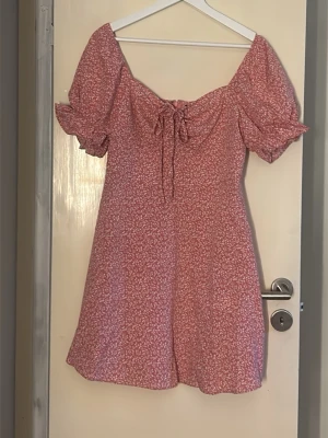 Rosa blommig kortklänning med puffärm - Söt rosa kortklänning med små vita blommor, puffärmar och knytning framtill. Klänningen har volangdetaljer vid ärmslut och en feminin, lätt utställd passform. Perfekt för varma dagar och somriga tillfällen. Endast testad