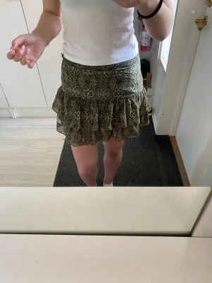 Grön mönstrad volangkjol - Supersöt kort kjol i grönt med svart mönster och volanger. Kjolen har en figurnära passform upptill och två lager volanger som ger en lekfull look. Perfekt att matcha med en enkel topp för en trendig outfit.