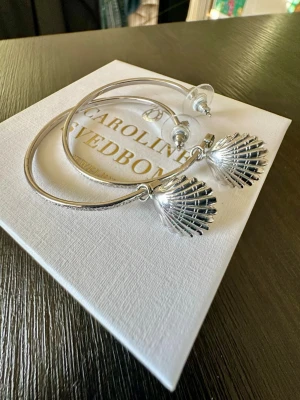 Caroline Svedbom Shell Loop Earrings - Stilrena örhängen i silverfärgad metall från Caroline Svedbom. De har stora runda hoops med hängande snäckformade berlocker som ger en cool och trendig look.