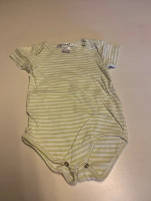 Randig kortärmad body från ECO BOO - Söt body med korta ärmar från ECO BOO i ljusgult och vitt randigt mönster. Bodyn har praktiska tryckknappar i grenen för enkel på- och avklädning. Tillverkad i mjukt material som är skönt mot huden.